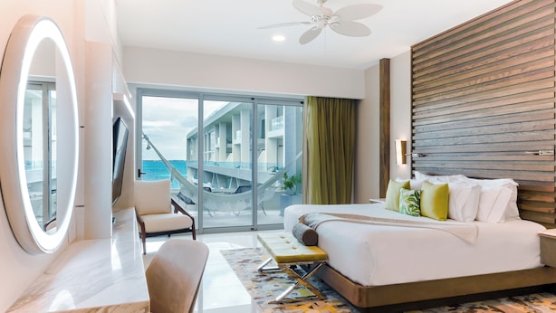 Una habitación de hotel moderna con una cama grande, un área de estar, un tocador con espejo y un balcón con vista al mar.