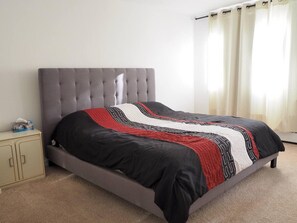 Ein Schlafzimmer mit einem grau gepolsterten Bett, einem beigen Schrank und einem Fenster mit Vorhängen.