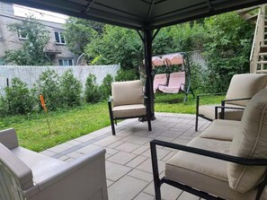 An outdoor patio area with two beige chairs and a swing chair, surrounded by a white fence and greenery. - Cozy Montreal Retreat - Shops - walk to everything (Montreal)