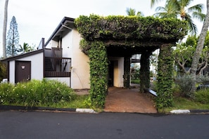 Ein Haus mit überdachter Veranda, einer kleinen Terrasse und einem Garten mit tropischen Pflanzen.