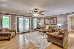 A spacious living room with a ceiling fan, a large window with curtains, a brown leather sofa, a coffee table, and a patterned rug. - Lake Wateree home with your own private dock and ramp.  Pet friendly with a fee. (Liberty Hill)