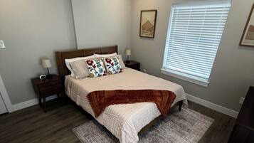 A bedroom with a bed, nightstands, a window with blinds, a rug, and a guitar.