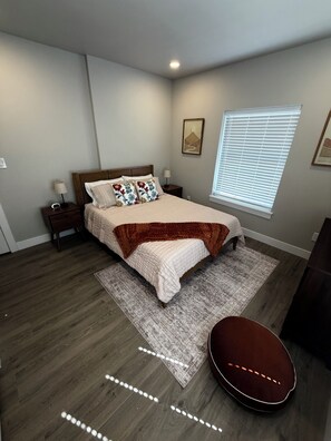 A bedroom with a bed, nightstands, a window with blinds, a rug, and a guitar.