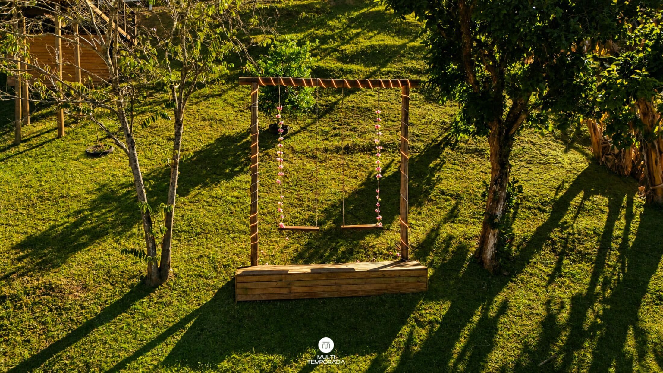 Un columpio de madera en un área con césped, árboles y una estructura de madera al fondo.