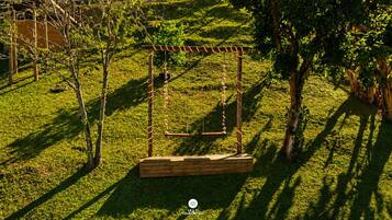 Un columpio de madera en un área con césped, árboles y una estructura de madera al fondo.