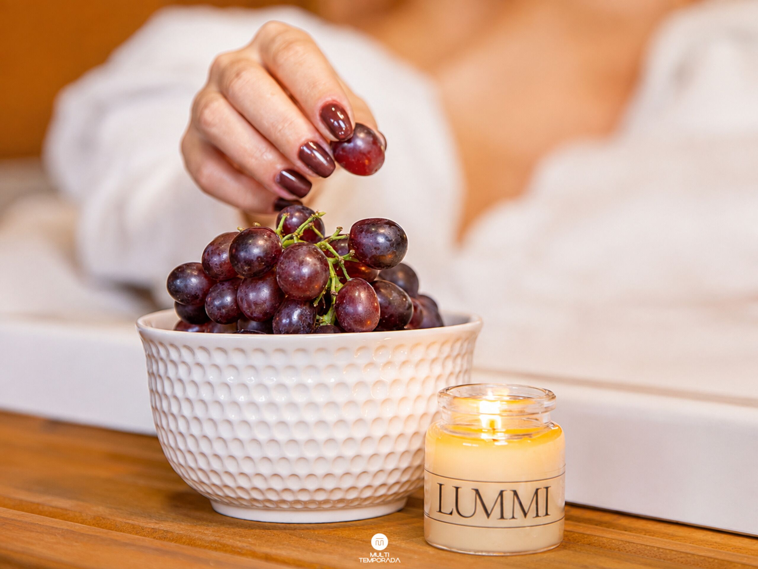 A white textured bowl filled with red grapes, a lit candle with 'LUMMI' on it, and a wooden surface.