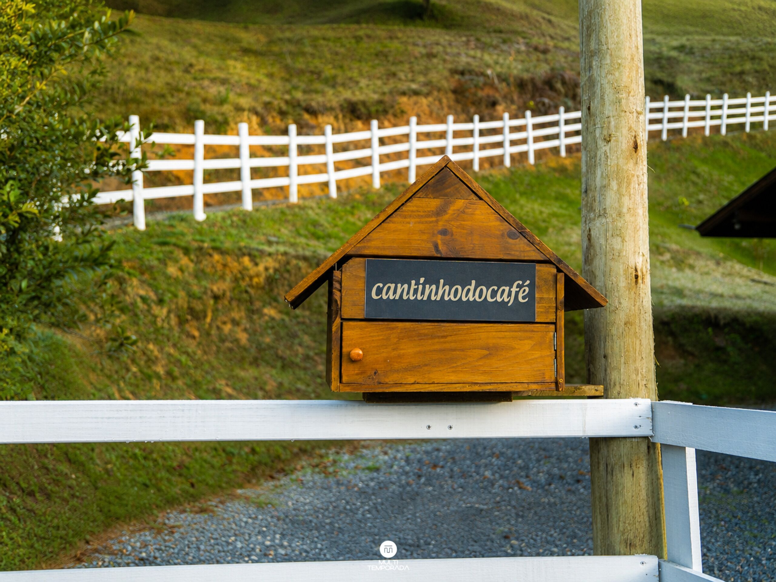 Ein hölzernes Schild mit dem Schriftzug 'cantinhodocafé' an einem weißen Zaun.