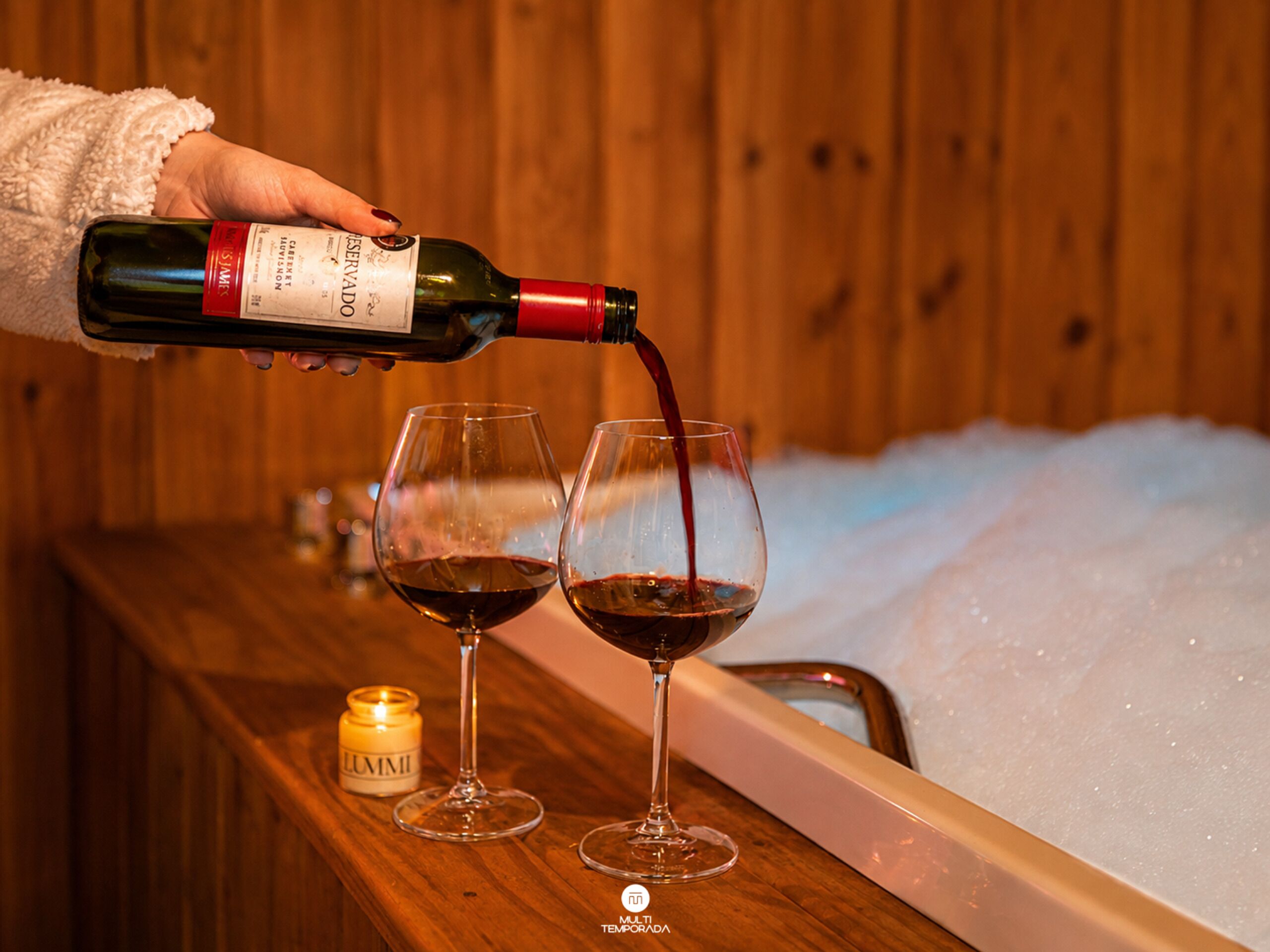 Une main verse du vin dans deux verres sur un comptoir en bois, avec une bougie et un seau à glace en arrière-plan.