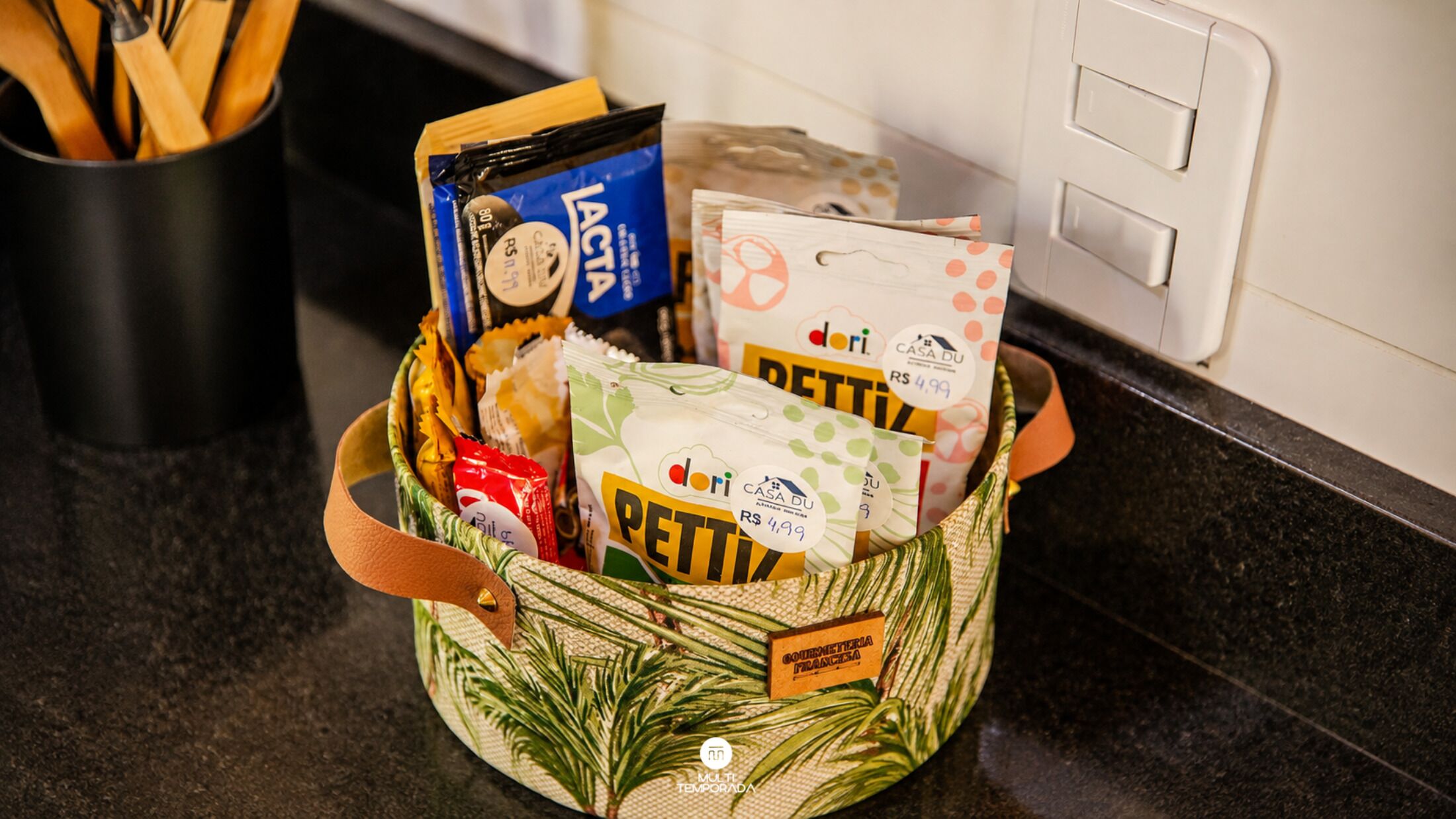 A kitchen counter with a basket containing various food items and a black container with wooden utensils.