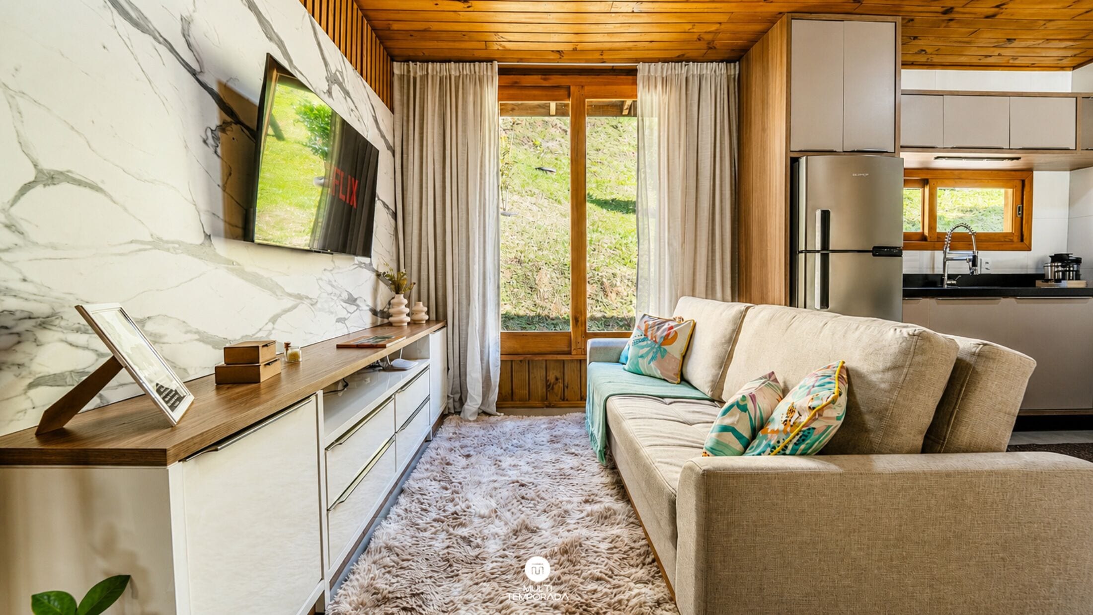 Una sala de estar acogedora con un sofá, un televisor montado en una pared de mármol, techo de madera y vistas al exterior a través de una ventana.