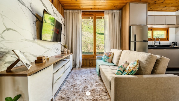 A cozy living area with a sofa, a TV mounted on a marble wall, a wooden ceiling, and a view of the outdoors through a window.