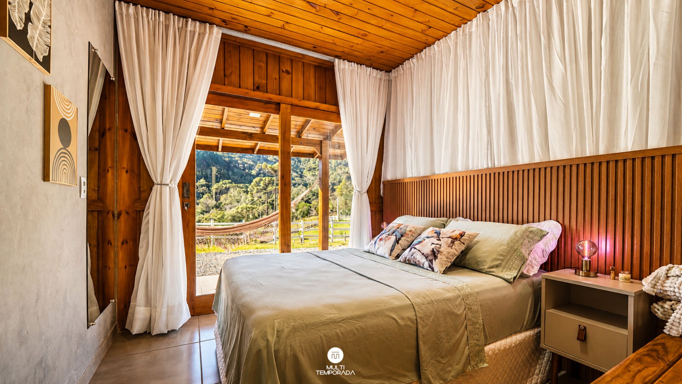 A bedroom with a wooden ceiling, a bed with a green bedspread, a nightstand with a lamp, and a view of a mountainous landscape through a large window.