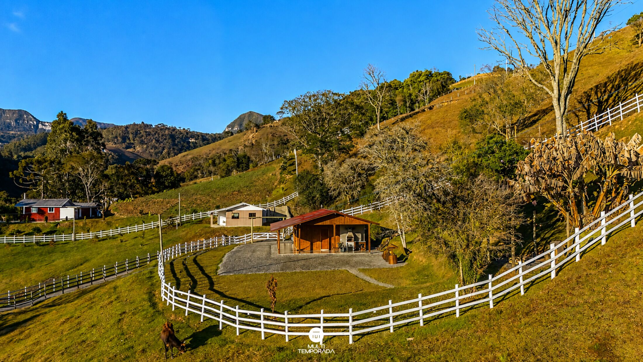 A fenced property with a red-roofed building, a white fence, and a hilly landscape.