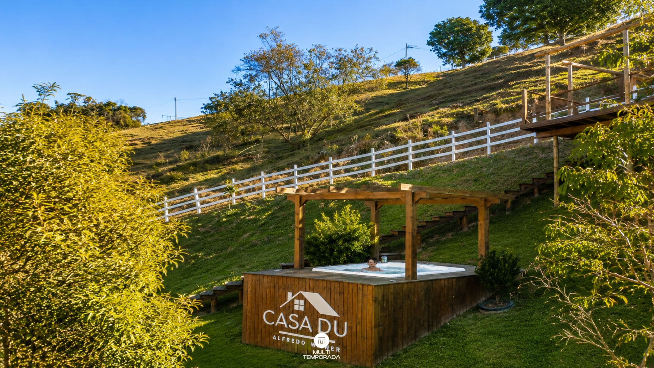 Una estructura de madera con un letrero que dice 'CASADU ALFREDO WILMER MULTI TEMPORADA' en una colina cubierta de hierba, con una cerca blanca y árboles al fondo.