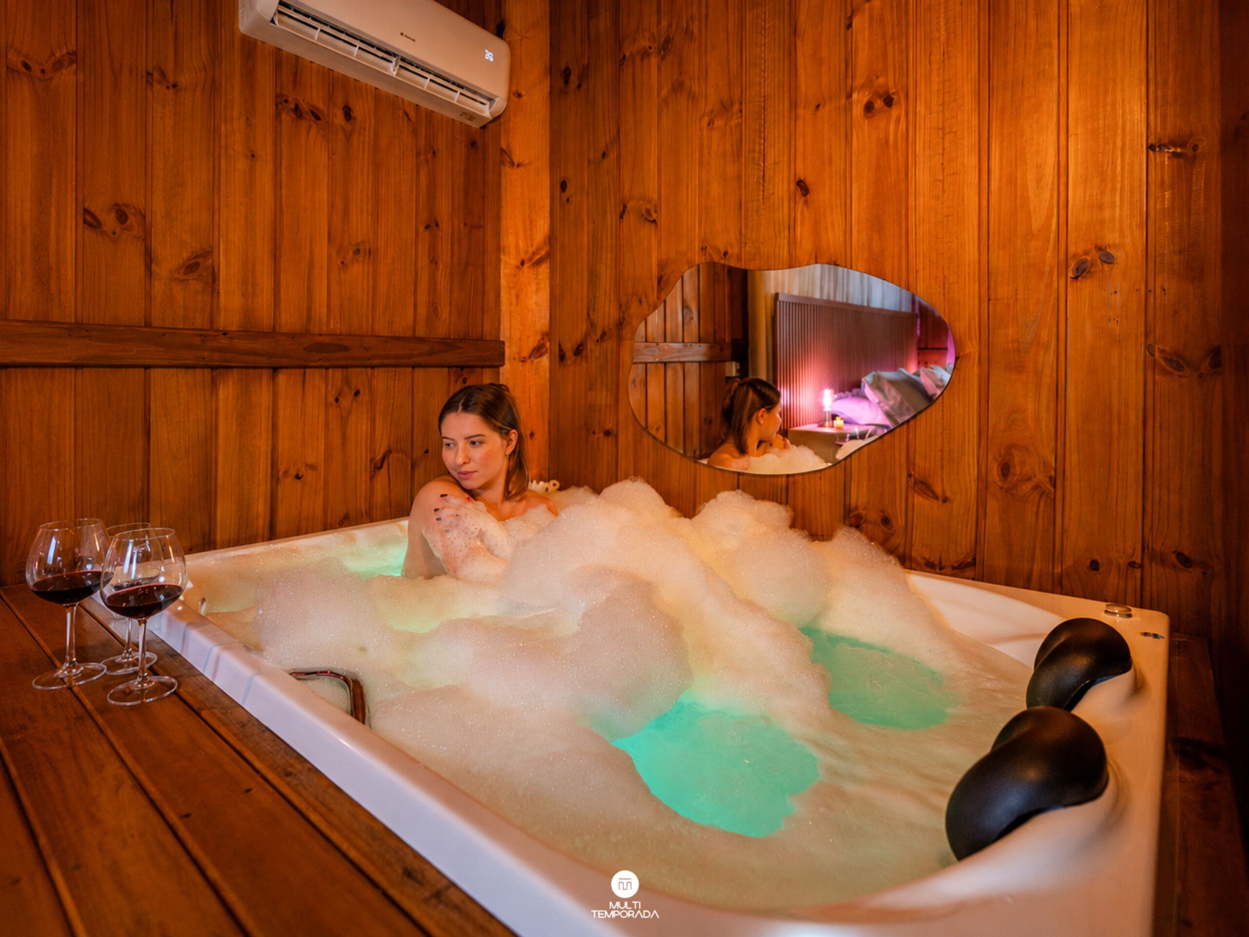 A wooden cabin interior with a bathtub filled with bubbles, a mirror, and three glasses of red wine on a wooden table.