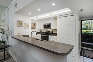 A modern kitchen with white cabinetry, a granite countertop, stainless steel appliances, and artwork on the walls. - Bonita Bay Lakefront Condo w/Private Beach and Pool (Bonita Springs)