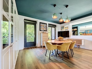 A dining area with a round table and chairs, a television, and a large window with a view of greenery.