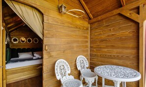 Interior de uma cabana de madeira com uma cama, duas cadeiras, uma mesa e um candeeiro pendente.