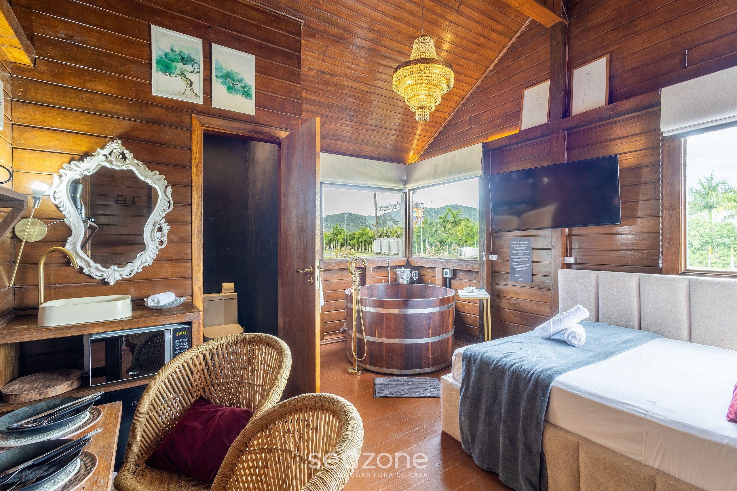 A wooden cabin interior with a bed, wicker chairs, a wooden bathtub, and a television.