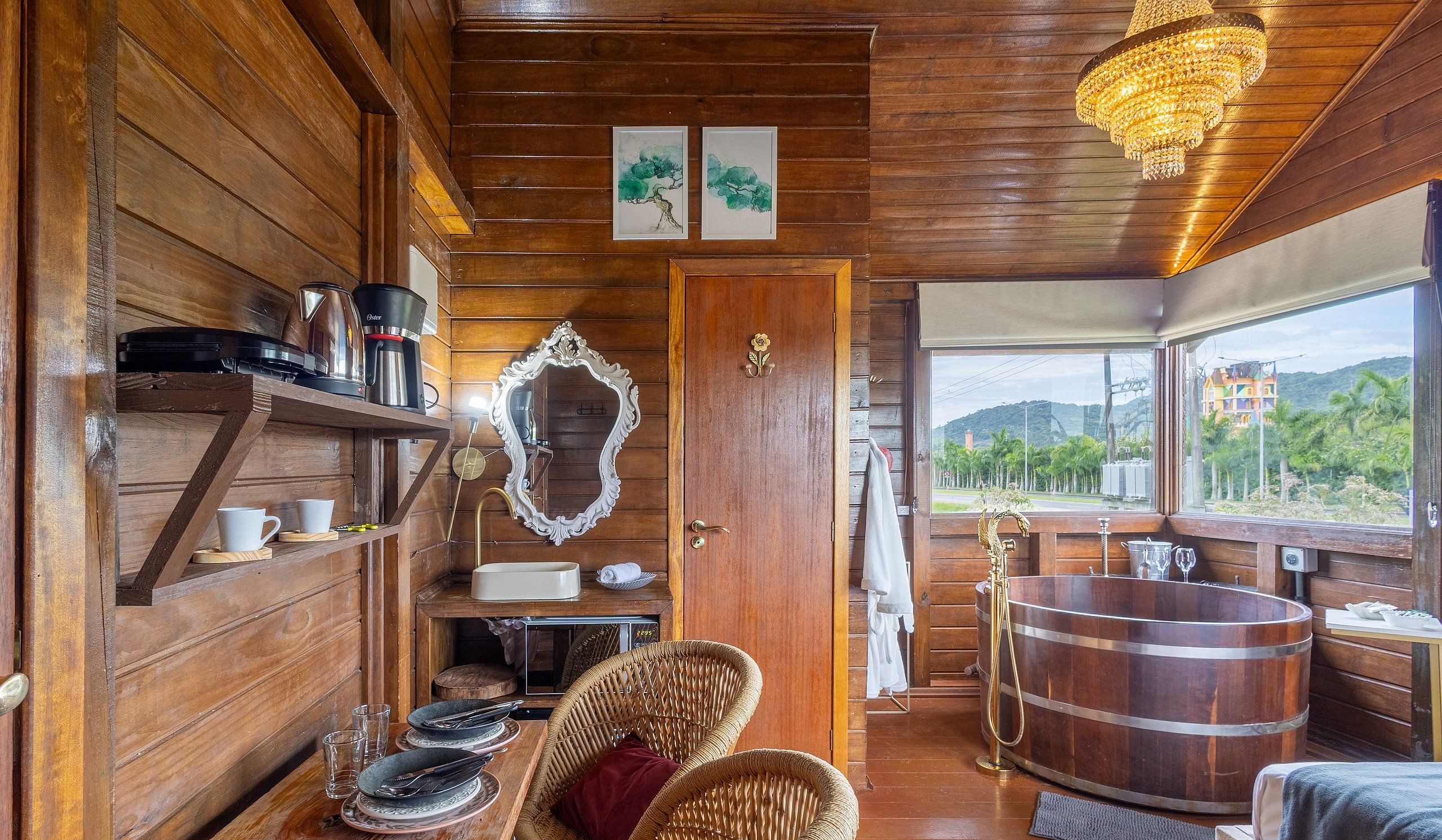 A wooden cabin interior with a dining area, a large wooden bathtub, and a view of a colorful building through a window.