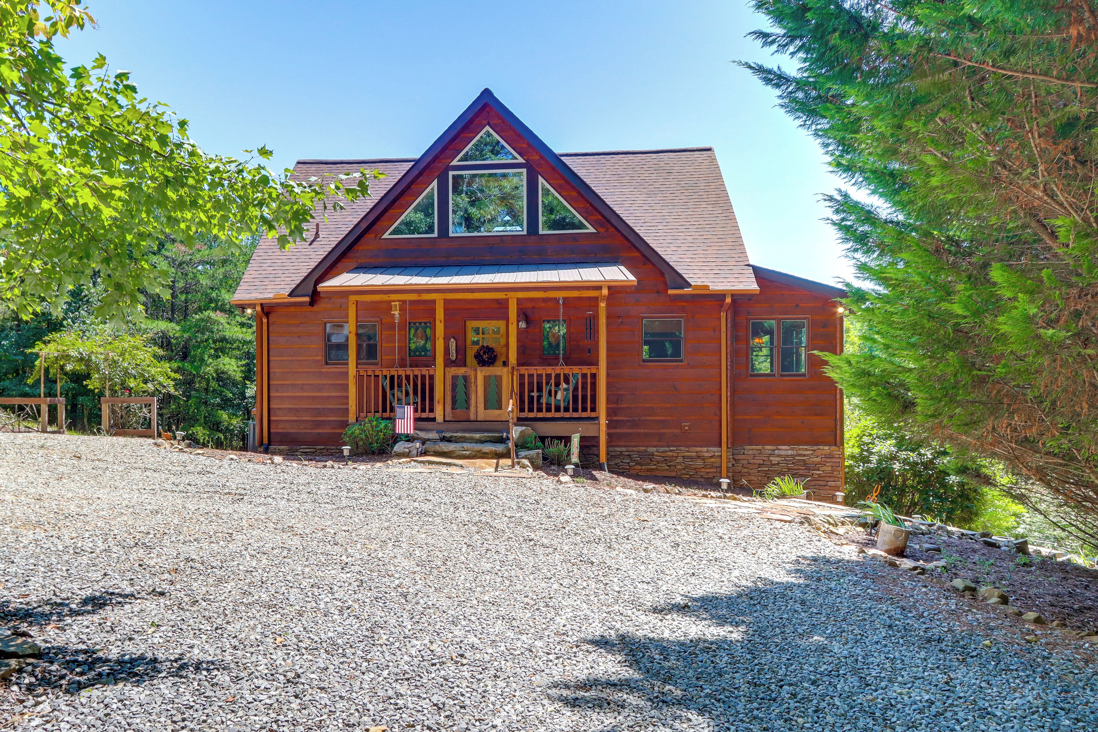 A wooden cabin with a porch, a stone foundation, and a gravel driveway.