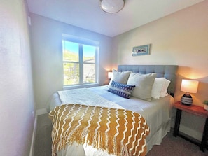 Une chambre à coucher avec un lit, une fenêtre, une table de chevet avec une lampe et un tableau accroché au mur.