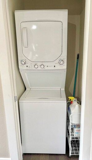 Une petite machine à laver blanche à chargement frontal, installée dans un petit local buanderie, avec un balai et des produits de nettoyage à proximité.