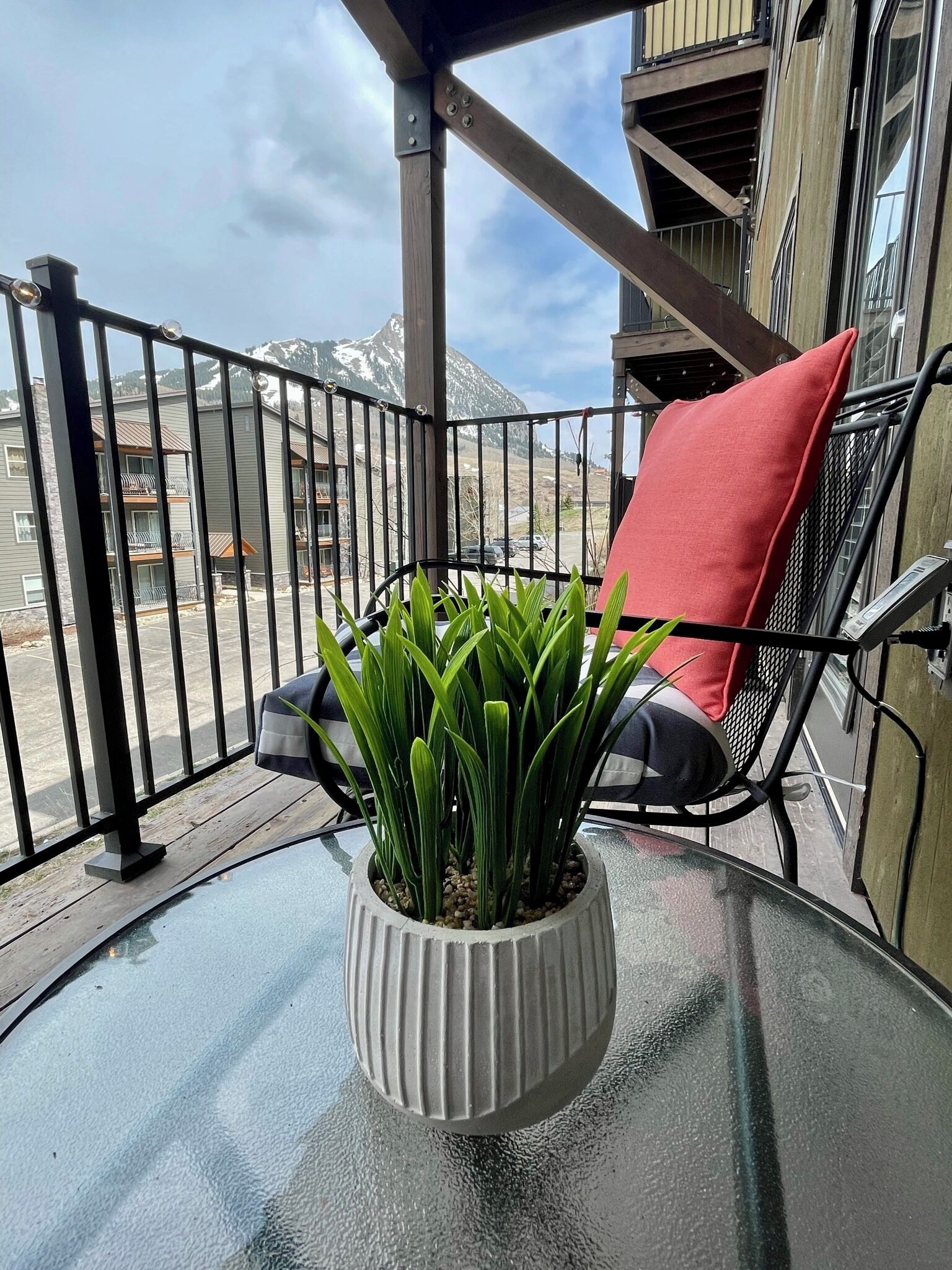 A balcony with a glass table, a potted plant, a red cushion, and a view of a mountain.