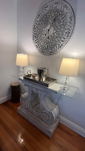 A room with a glass console table, two lamps, a decorative clock, and a trash can.