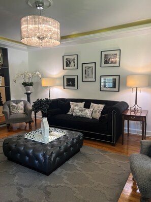 A living room with a black sofa, a white ottoman, a chandelier, and framed pictures on the wall.