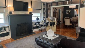 A living room with a fireplace, a television, and a chandelier. - Charming 3BR cottage, that borders the grounds of Historic Linden. (Natchez)