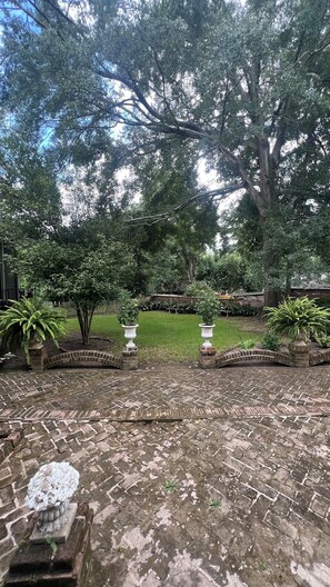 Une terrasse en briques avec une fontaine, entourée d’une végétation luxuriante et d’arbres.