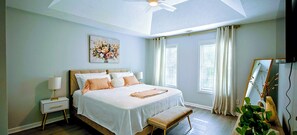A bedroom with a bed, bedside table, lamp, curtains, windows, and a painting on the wall.