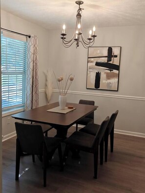 A dining area with a rectangular table, six chairs, a chandelier, a vase with dried flowers, and a painting on the wall.