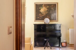 A room with a black piano, a framed painting, a small stool, and a radiator. - Gemma apartment in Venice (Venezia)
