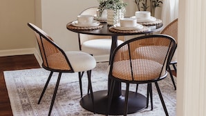 A dining area with a round table, four chairs, a hanging light fixture, a framed artwork, and a potted plant.