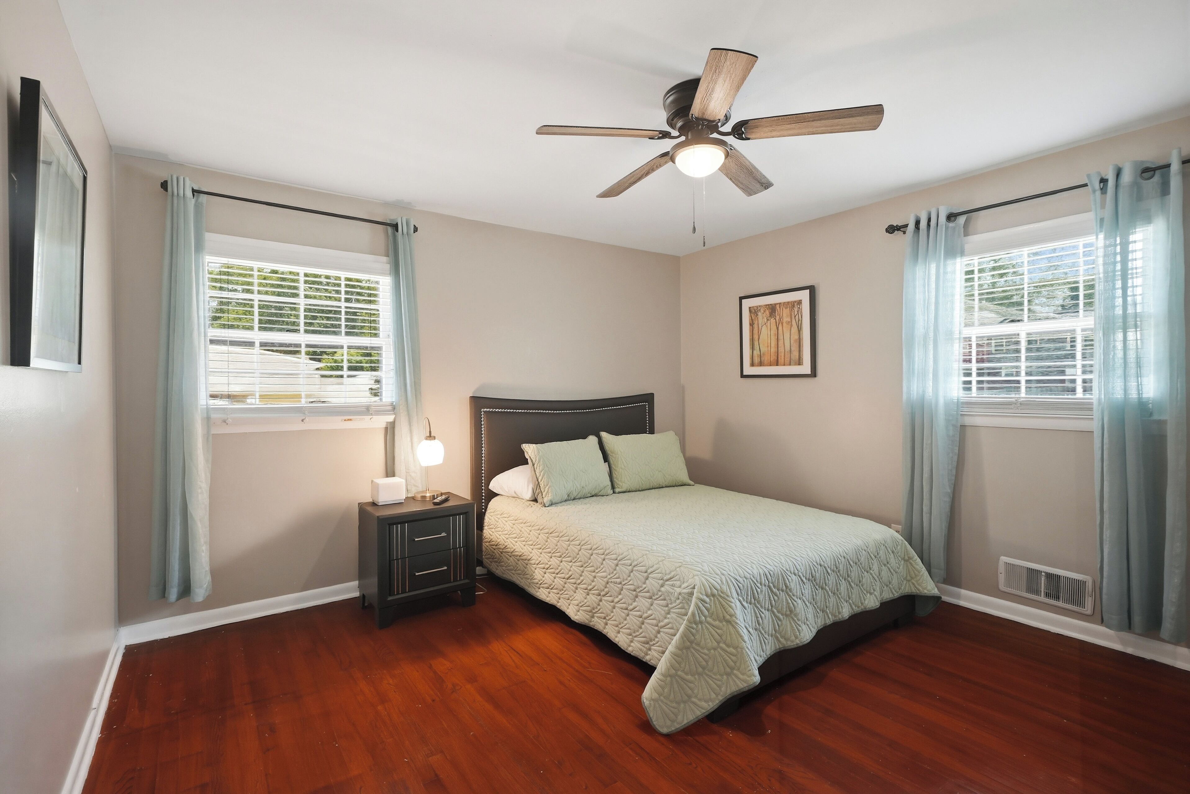 Une chambre à coucher avec un lit, une table de chevet, un ventilateur de plafond et des fenêtres avec des rideaux.