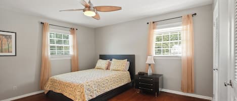 A bedroom with a bed, nightstand, lamp, ceiling fan, and windows with curtains.