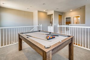 Mesa de bilhar com bolas e tacos, cercada por uma proteção branca, em um ambiente com cores neutras e iluminação embutida.