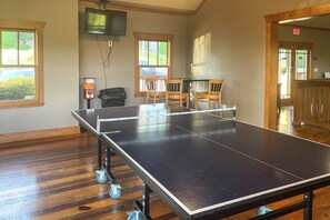 Una mesa de ping pong con ruedas, un televisor fijado a la pared y un ventanal con vistas a un coche y follaje.