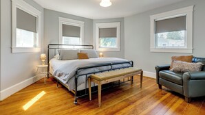 Ein Schlafzimmer mit einem Bett, Nachttischen, einer Bank, einem Sessel und großen Fenstern mit Jalousien.