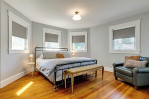Un dormitorio con una cama, mesitas de noche, un banco, una silla y ventanas amplias con persianas.