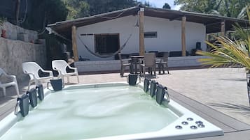 A hot tub with chairs and tables, a hammock, and a building with a stone wall and wooden pillars.