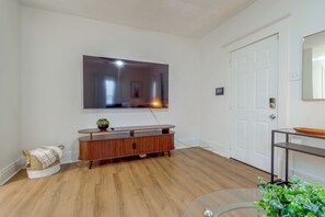 A modern living room with a flat-screen TV mounted on the wall, a wooden entertainment unit, a glass coffee table, and a door with a mirror.