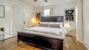 Quarto com cama de madeira escura, ventilador de teto, mesa de cabeceira com abajur e obras de arte nas paredes.