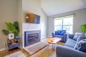 Een woonkamer met een open haard, een flatscreen televisie erboven, een bank, een salontafel en een raam met uitzicht op bomen.