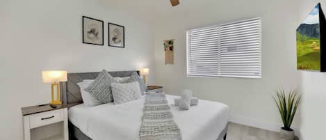 A bedroom with a bed, nightstands, a ceiling fan, a window with blinds, and a wall-mounted picture.