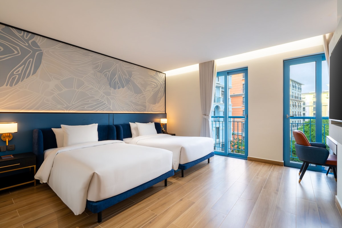 Habitación de hotel con dos camas, un cuadro abstracto grande en la pared, un balcón con vistas a la ciudad y un escritorio con una silla.