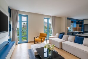 Ein modernes Wohnzimmer mit einer Couch, einem Couchtisch, einem Fernseher und einem Sessel. Große Fenster mit Vorhängen, eine Küchenzeile und ein Balkon mit Blick auf Palmen.