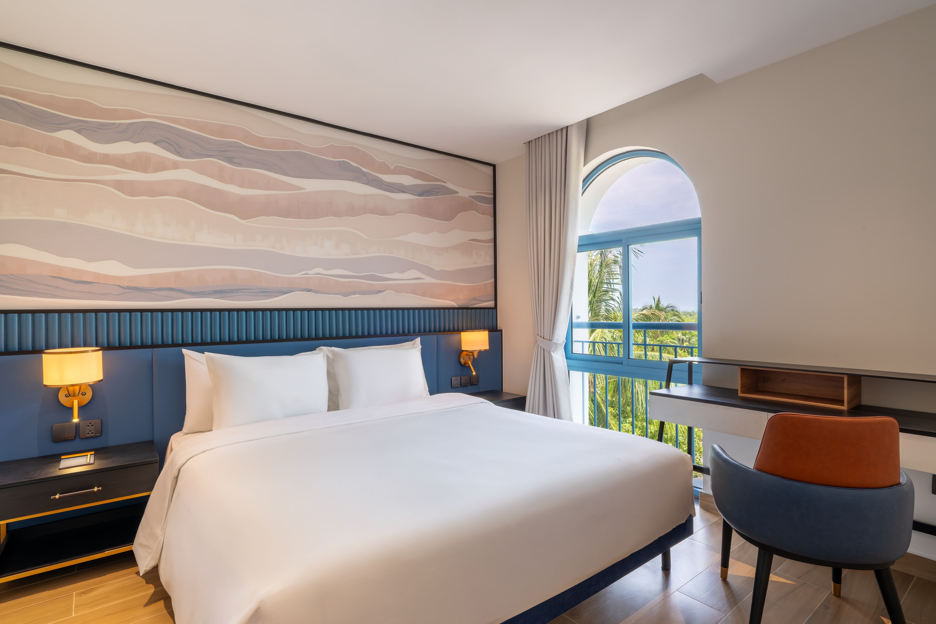 Habitación de hotel con una cama grande, dos lámparas de noche, un escritorio con una silla y vistas a zonas verdes por una ventana.